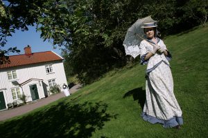 Kvinna står utklädd på gräsmattan framför det vita trähuset Lilla Änggården