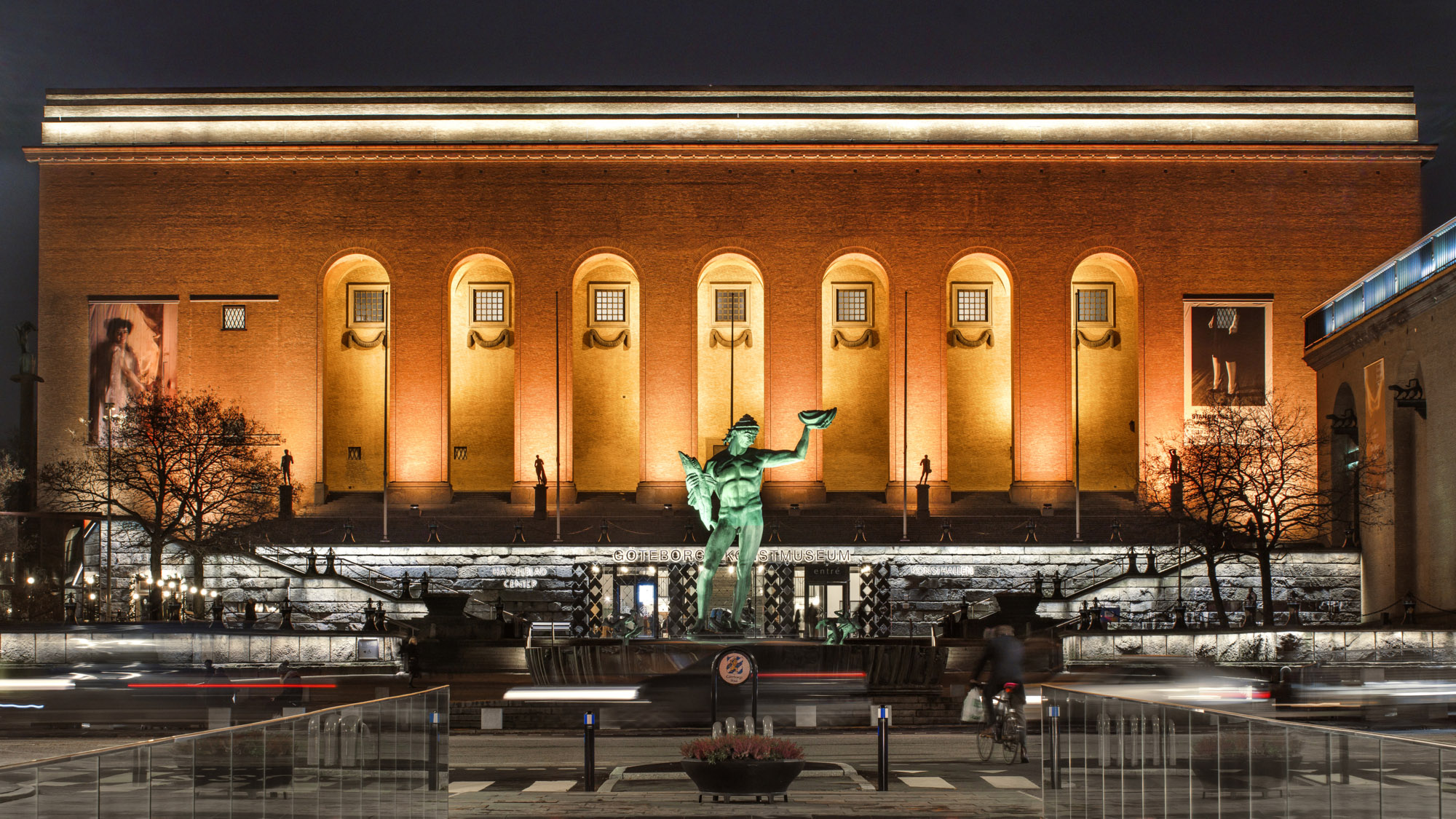 Kvällsbild på upplyst byggnad med valvbågar och staty i förgrunden